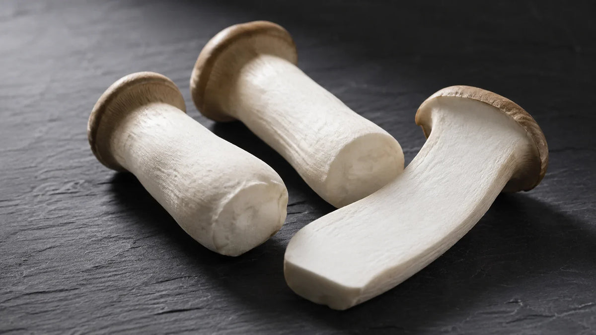 Three fresh king oyster mushrooms on dark slate, one halved lengthways to show the dense white stem interior.