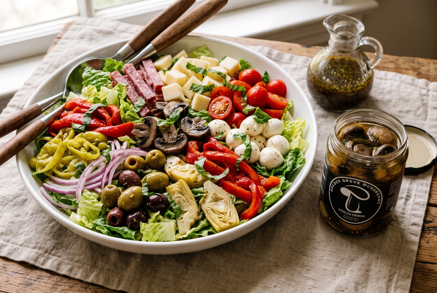 A bowl of antipasto salad with romaine, salami, provolone, mozzarella balls, tomatoes, roasted peppers, artichokes, olives, pepperoncini and pickled crimini, beside an open jar of Golden Grove pickled crimini