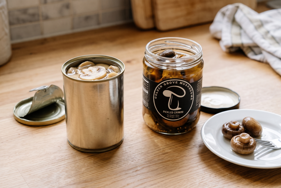An open can of sliced mushrooms next to an open jar of Golden Grove pickled crimini, with three whole pickled crimini caps and a fork on a small plate