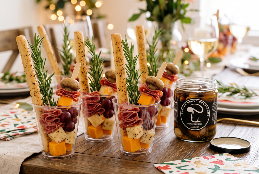 Clear charcuterie cups with breadsticks, rosemary, cheese cubes, folded salami, grapes and skewers topped with pickled crimini, beside an open jar of Golden Grove pickled crimini