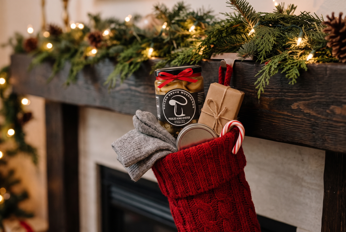 A red knit Christmas stocking on a mantel holding a jar of Golden Grove pickled king oyster tied with a red ribbon, wool socks, a small wrapped box, a tin and a candy cane