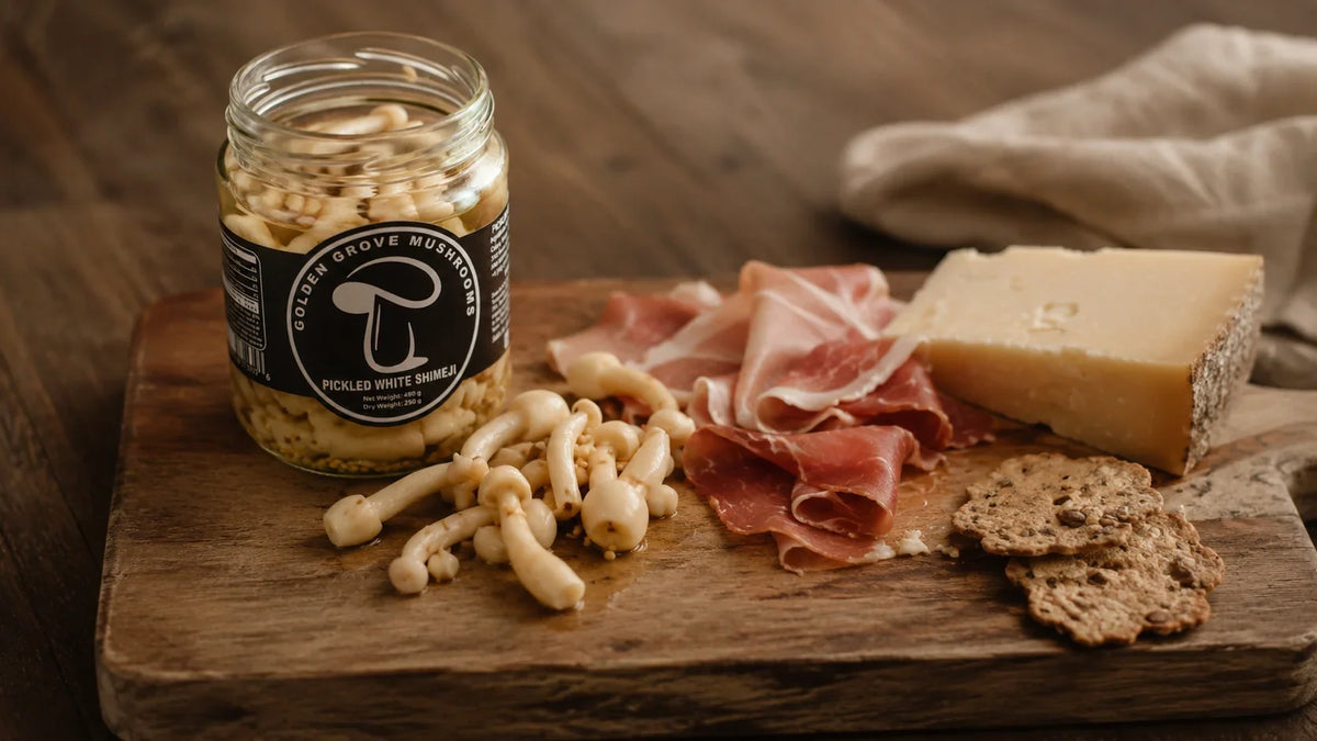 An open jar of pickled white shimeji mushrooms on a wooden board with prosciutto, aged hard cheese and seeded crackers.
