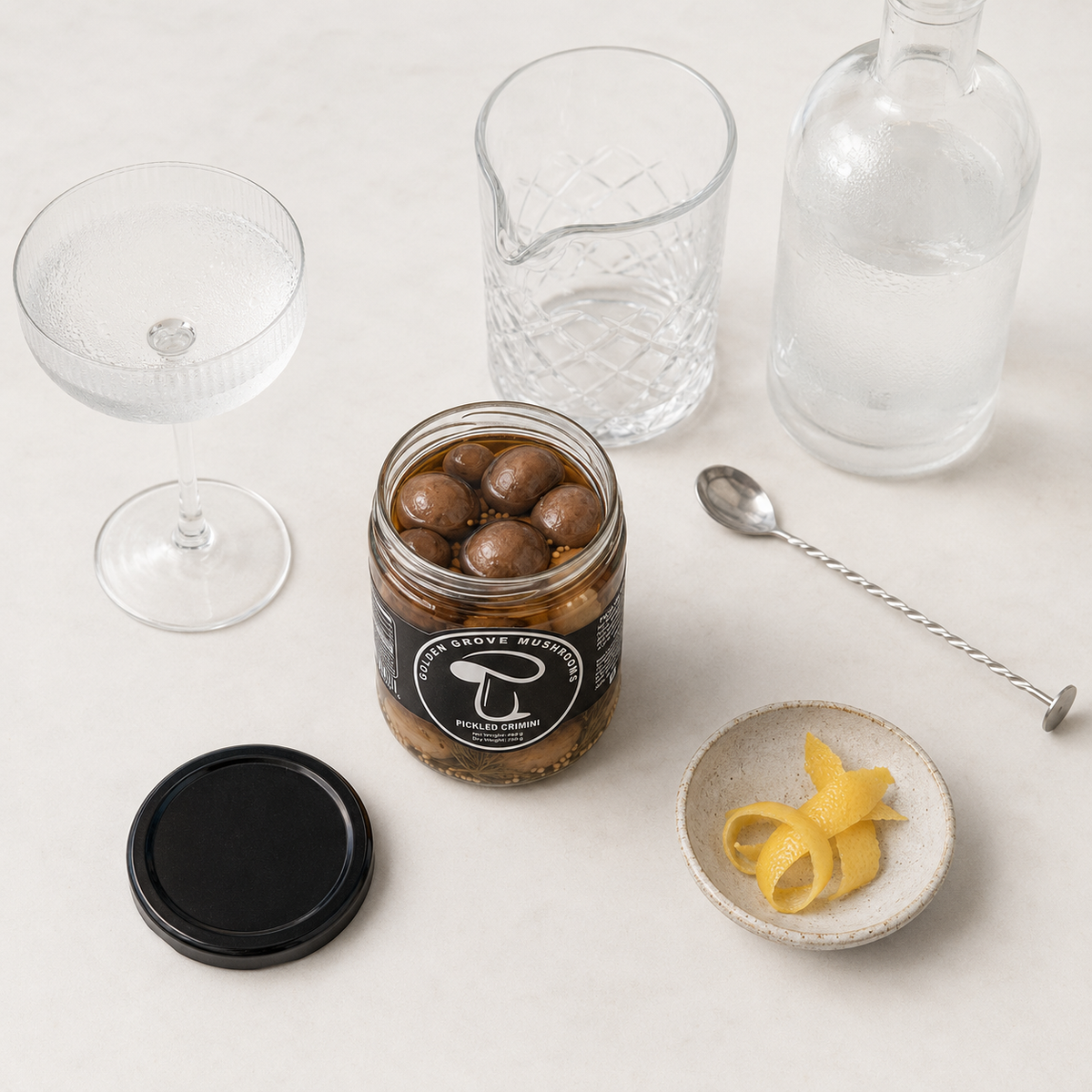 Coupe glass, mixing glass, bar spoon and a jar of pickled crimini laid out on a bar counter