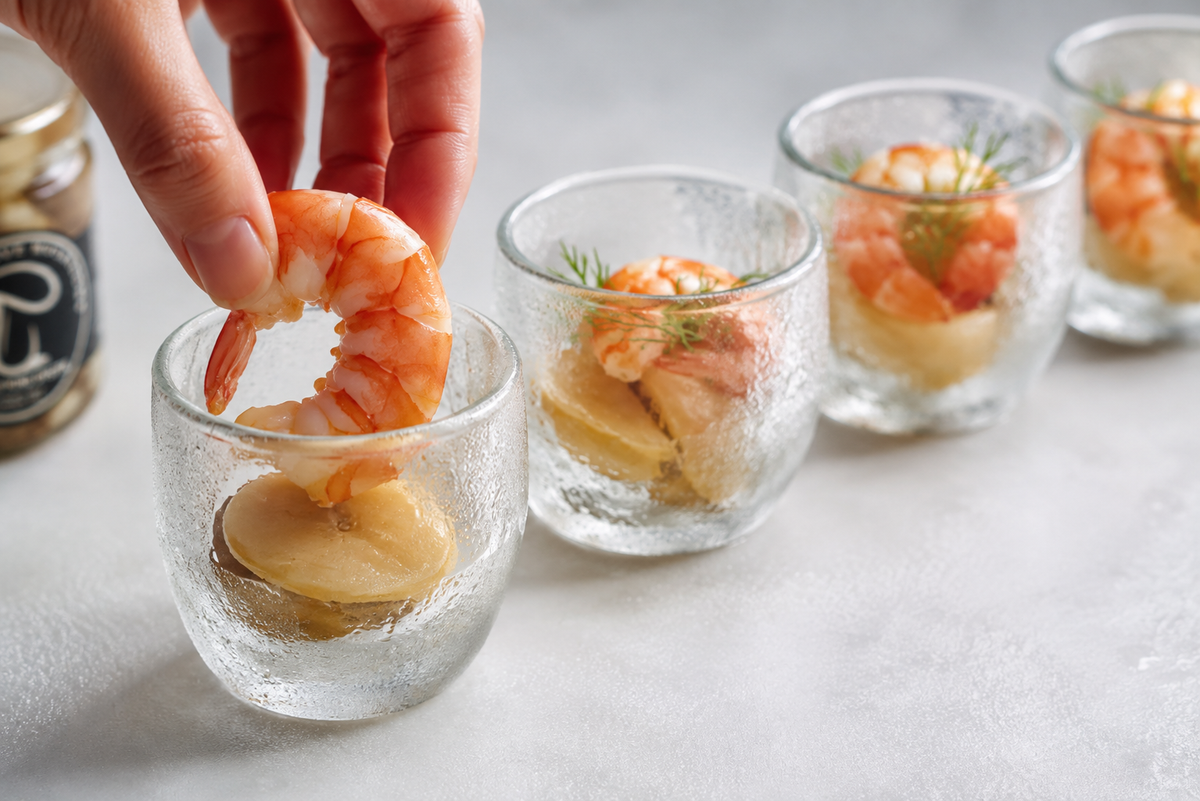 A hand laying a poached prawn into a chilled glass cup holding king oyster coins