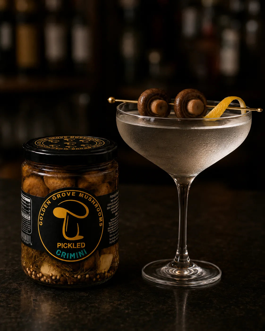 A coupe glass martini garnished with two pickled crimini mushrooms and a lemon twist on a gold pick, beside a jar of pickled crimini on a dark bar.